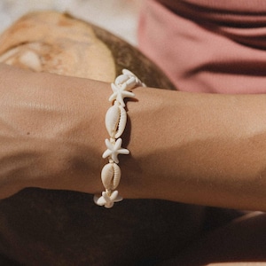 May include: A cream-colored bracelet featuring alternating seashells and starfish. The bracelet is made of natural materials and is worn on a person's arm. The bracelet is a beach-themed accessory.
