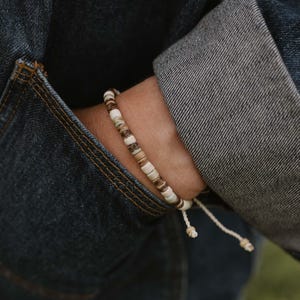 Peut inclure: Un bracelet de perles alternant des perles marron clair et blanches. Le bracelet est monté sur un cordon crème avec des nœuds réglables. Le bracelet est porté sur un poignet, avec une veste en jean en arrière-plan.