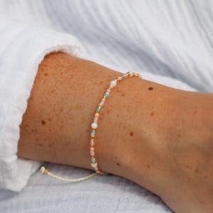 Pode incluir: Uma delicada pulseira de contas com uma mistura de contas em tons pastel, incluindo rosa, azul e branco, intercaladas com contas douradas. A pulseira é enfiada em um cordão creme e é mostrada em um pulso.