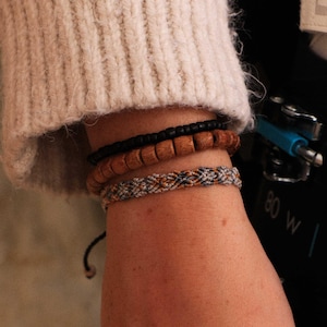 Pode incluir: Close-up de três pulseiras empilhadas em um pulso. A pulseira superior é preta, a do meio é feita de contas de madeira e a inferior é um estilo macramê tecido em tons de cinza, laranja e preto. A pessoa está usando um suéter creme.