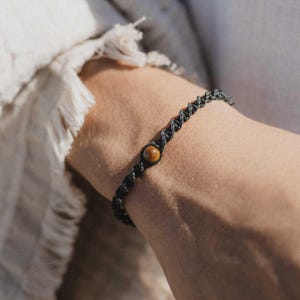 May include: A black macrame bracelet with a single, round, brown tiger's eye bead. The bracelet is worn on a wrist, with a soft, off-white fabric in the background. The macrame design is visible, showcasing the intricate knotting.