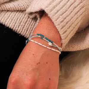 May include: Three beaded bracelets in shades of white, turquoise, and blue. One bracelet features a white star charm. The bracelets are worn on a wrist, with a cream-colored sweater in the background.