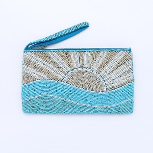 May include: A rectangular beaded clutch with a wrist strap. The design features a sun rising over a turquoise ocean. The sun is beige and white, and the waves are turquoise. The wrist strap is turquoise.