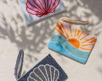 May include: Three beaded clutch bags. One is light blue with a pink and red seashell design. Another features a sunrise over blue waves. The third is navy blue with a gray and white seashell design.