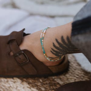 May include: Ankle bracelet with turquoise, white, and gold beads. The beaded bracelet is layered and worn on an ankle with a fern tattoo. A brown leather sandal is in the foreground.