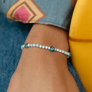 Może przedstawiać: Bransoletka z białych i niebieskich koralików z turkusowymi zawieszkami w kształcie tureckiego oka. Bransoletka jest na czyimś nadgarstku.