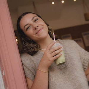 May include: A woman wearing a beige sweater and a gold bracelet holds a green smoothie in a clear plastic cup with a pink and white striped straw.