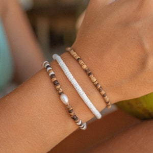 Puede incluir: Tres pulseras hechas de madera y cuentas blancas. La pulsera de abajo tiene una sola perla.