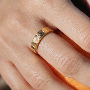 Könnte beinhalten: Ein goldfarbener Ring mit einem sich wiederholenden Sonnenmuster. Der Ring wird an einem Finger getragen, und das Sonnenmuster ist um das gesamte Band geätzt. Das Design des Rings ist detailliert und aufwendig, mit einer polierten Oberfläche.