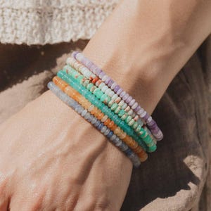 Könnte beinhalten: Eine Sammlung von Armbändern mit Perlen in verschiedenen Farben, darunter Lila, Grün, Orange und Blau. Die Armbänder bestehen aus kleinen, runden Perlen und sind an einem Handgelenk aufgereiht. Der Hintergrund ist ein neutrales Beige.