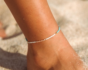 May include: A delicate anklet with white, turquoise, and gold beads. The anklet is made of small, evenly spaced beads and is a simple, elegant piece of jewelry.