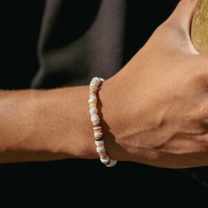 May include: A beaded bracelet featuring a mix of white, tan, and brown disc-shaped beads. The bracelet is strung together, creating a simple yet stylish accessory. The bracelet is worn on a wrist against a dark background.