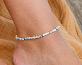 May include: A beaded anklet with blue, white, and beige beads. The anklet is on a person's ankle.