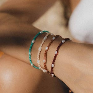 May include: Four macrame bracelets in teal, cream, brown, and burgundy, each decorated with small, white pearls. The bracelets are displayed on an arm, showcasing the different colours and textures of the jewellery.