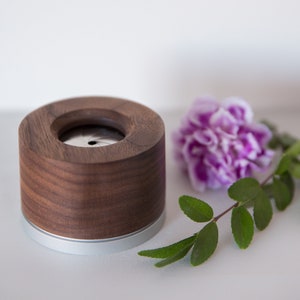 May include: A wooden and metal incense holder with a silver band around the bottom. The holder is sitting on a white surface next to a purple flower and green leaves.