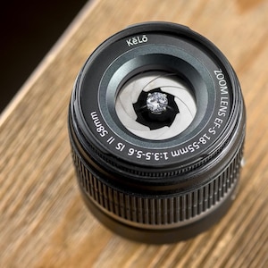 Puede incluir: Un anillo de compromiso de diamantes está centrado dentro de un objetivo de cámara negro. La lente tiene el texto "Kēlo" y "ZOOM LENS EF-S 18-55mm 1:3.5-5.6 IS II 58mm" impreso. La lente está sobre una superficie de madera.