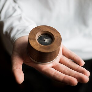 May include: A small, round, wooden box with a silver band around the top. The box has a circular opening on the top that reveals a black, multi-bladed shutter.