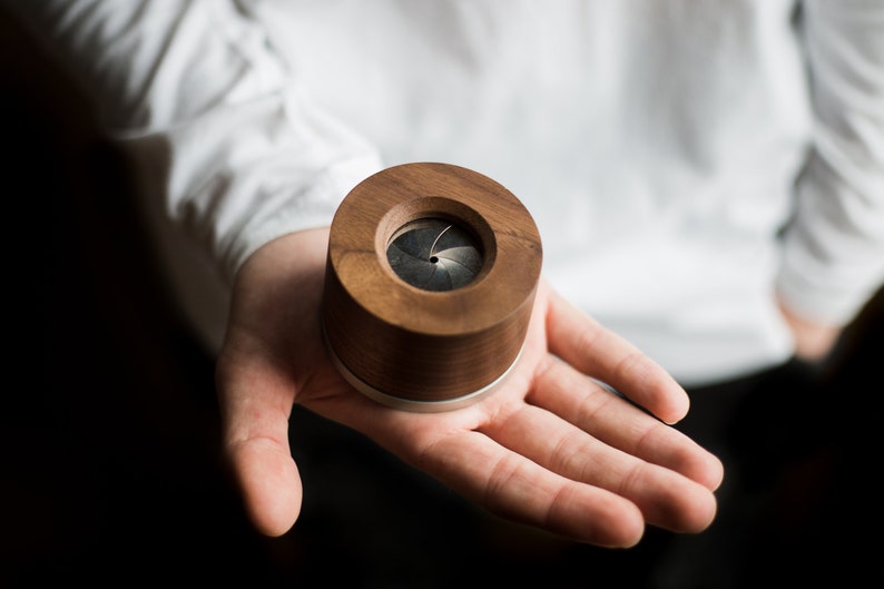May include: A small, round, wooden box with a silver band around the top. The box has a circular opening on the top that reveals a black, multi-bladed shutter.