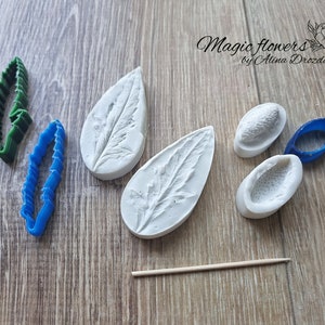 May include: A set of six silicone molds for making leaf and flower shapes. The molds are white and have a textured surface. There are two leaf molds, two flower bud molds, and two small round molds. The molds are arranged on a wooden surface.  Magic Flowers by Alina Drozdlova.