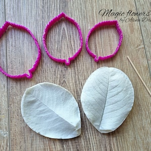 Può includere: Tre tagliapasta per fiori in plastica rosa di diverse dimensioni, due forme di foglie bianche e uno spiedino di legno su una superficie in legno. Magic flowers by Alina Drezdova.