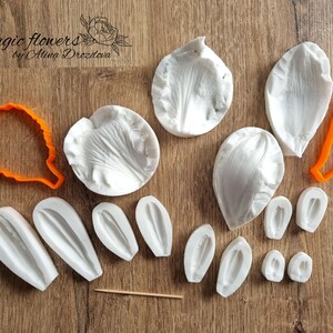 Op de afbeelding: Een set siliconen mallen voor het maken van bloemblaadjes. De mallen zijn wit en zijn verkrijgbaar in verschillende maten en vormen. De set bevat ook twee oranje plastic uitstekers. De tekst "Magic flowers by Alina Drozdova" is zichtbaar in de linkerbovenhoek van de afbeelding.