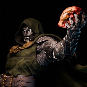 Puede incluir: Figura de acción detallada de Doctor Doom, vestido con una capa con capucha verde y armadura metálica. La figura sostiene una cabeza cortada y ensangrentada en su mano derecha, con una máscara plateada y detalles dorados.