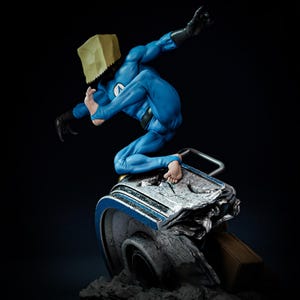 May include: A blue superhero figure in a dynamic pose, wearing a blue suit, black gloves, and a paper bag over the head. The figure is perched on a metallic, damaged object with a blue stripe. The background is dark.