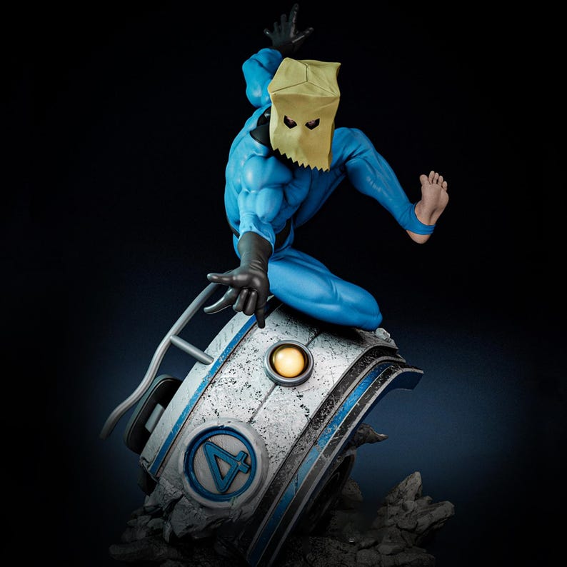 May include: A blue action figure of a superhero wearing a paper bag mask, black gloves, and boots. The figure is posed on a futuristic, metallic base with the Fantastic Four logo. The base has a light and is surrounded by rock-like structures.