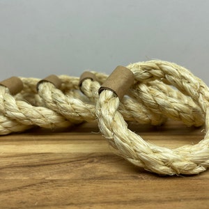 Puede incluir: Un conjunto de cinco servilleteros de cuerda trenzada. Los anillos est&aacute;n hechos de cuerda gruesa de color natural, con una banda de papel marr&oacute;n que asegura el cierre del anillo. Los anillos est&aacute;n dispuestos sobre una superficie de madera.