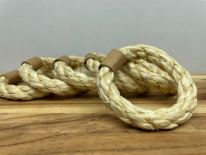 Puede incluir: Un conjunto de cinco servilleteros de cuerda trenzada. Los anillos est&aacute;n hechos de cuerda gruesa de color natural, con una banda de papel marr&oacute;n que asegura el cierre del anillo. Los anillos est&aacute;n dispuestos sobre una superficie de madera.