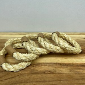 Puede incluir: Cuatro servilleteros de cuerda natural con detalles de papel marr&oacute;n. Los anillos est&aacute;n hechos de cuerda trenzada gruesa y est&aacute;n dispuestos sobre una superficie de madera. La paleta de colores neutros y la textura sugieren un tema r&uacute;stico o n&aacute;utico.