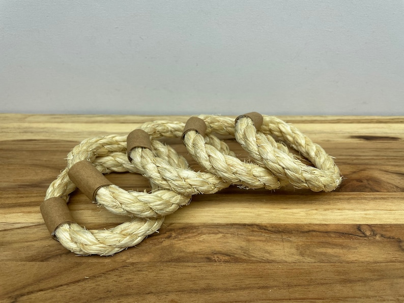 Puede incluir: Cuatro servilleteros de cuerda natural con detalles de papel marr&oacute;n. Los anillos est&aacute;n hechos de cuerda trenzada gruesa y est&aacute;n dispuestos sobre una superficie de madera. La paleta de colores neutros y la textura sugieren un tema r&uacute;stico o n&aacute;utico.