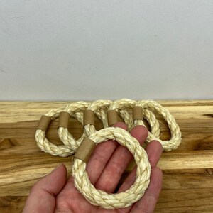 Puede incluir: Un conjunto de seis servilleteros de cuerda natural. Cada anillo est&aacute; hecho de cuerda de color blanquecino, bien enrollada, con una peque&ntilde;a banda de papel marr&oacute;n. Los anillos est&aacute;n sobre una superficie de madera.