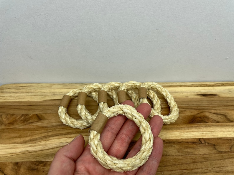 Puede incluir: Un conjunto de seis servilleteros de cuerda natural. Cada anillo est&aacute; hecho de cuerda de color blanquecino, bien enrollada, con una peque&ntilde;a banda de papel marr&oacute;n. Los anillos est&aacute;n sobre una superficie de madera.