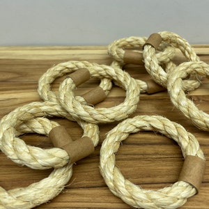 Puede incluir: Un conjunto de servilleteros de cuerda natural. Cada anillo est&aacute; hecho de cuerda gruesa y tejida en color beige claro. Los extremos de cada anillo est&aacute;n envueltos con una banda de papel marr&oacute;n. Los anillos est&aacute;n dispuestos sobre una superficie de madera.