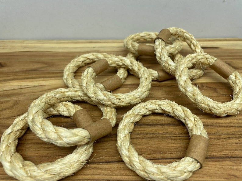 Puede incluir: Un conjunto de servilleteros de cuerda natural. Cada anillo est&aacute; hecho de cuerda gruesa y tejida en color beige claro. Los extremos de cada anillo est&aacute;n envueltos con una banda de papel marr&oacute;n. Los anillos est&aacute;n dispuestos sobre una superficie de madera.