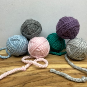 May include: Six balls of yarn in various colors, including gray, light blue, pink, green, and purple. The yarn is arranged on a wooden surface.