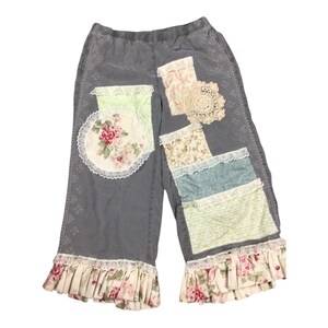 May include: Gray capri pants with floral and lace patchwork details. The pants feature a variety of fabric patches in pastel colors, including floral prints and lace trim. The hemline is finished with a ruffled floral fabric.
