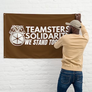 May include: A brown banner with white text that reads "Teamsters Solidarity We Stand Together". The banner features a white logo with two horses pulling a wheel with the words "International Brotherhood of Teamsters" around it. A person is hanging the banner on a white brick wall.