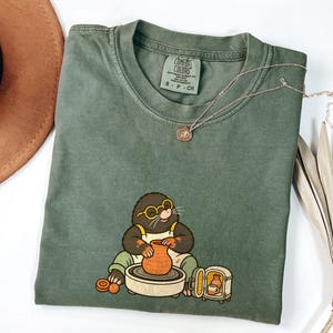 May include: An olive green short-sleeve t-shirt featuring a graphic of a mole wearing yellow glasses and an apron, working at a pottery wheel with a small kiln nearby. The shirt has a Comfort Colors brand tag at the neck and a crew neckline.