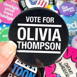 May include: A black button with the text "VOTE FOR OLIVIA THOMPSON" in white. The button is held in a hand and surrounded by other colorful buttons with various designs and text, including "design your own badge".