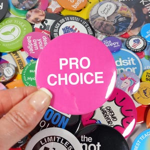 Puede incluir: Un botón rosa con el texto "PRO CHOICE" en letras blancas. El botón está rodeado de otros botones con varios diseños y textos.