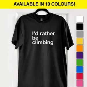 Op de afbeelding: Zwart T-shirt met de witte tekst "I'd rather be climbing". Het shirt hangt aan een zwarte hanger tegen een witte deur. Een gele banner bovenaan zegt "AVAILABLE IN 10 COLOURS!" met een kleurenkaart rechts.