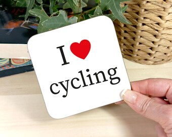 May include: A white square coaster with the words "I ❤️ cycling" in black and a red heart symbol. The coaster is held by a person's hand. The background includes a wicker basket and greenery.