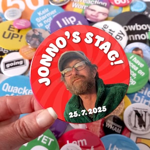 May include: A circular pin-back button featuring a red background with a swirling pattern. The button displays a photograph of a person with a beard wearing a cap and glasses, the text JONNO'S STAG! in white, and the date 25.7.2025 in white.