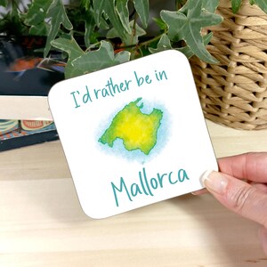 Puede incluir: Posavasos cuadrado blanco con el texto "I'd rather be in Mallorca" en color turquesa. El posavasos presenta un mapa de Mallorca en acuarela en verde y amarillo, rodeado de un lavado azul.