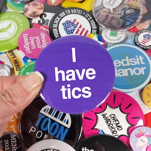 May include: A collection of colorful button pins with various designs and messages. One button pin is purple with white text that reads "I have tics".