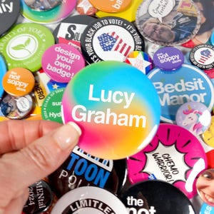 Op de afbeelding: Een verzameling kleurrijke knopen met verschillende ontwerpen en tekst. De knopen hebben een regenboog-tie-dye-ontwerp met de tekst "Lucy Graham" in wit.