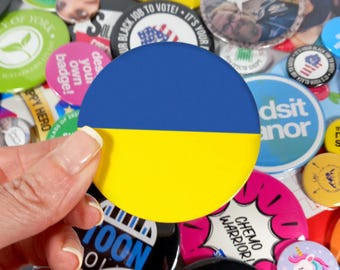 Könnte beinhalten: Ein blauer und gelber Ansteckbutton mit den Farben der ukrainischen Flagge. Der Button wird von einer Hand gehalten.