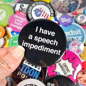 May include: A collection of colorful button pins with various designs and messages. One button pin features black text on a black background that reads "I have a speech impediment."